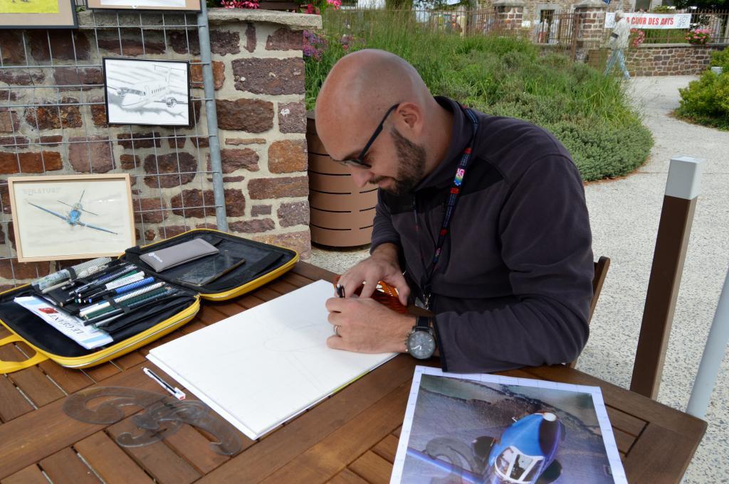 Clément Houdmond en train de réaliser un dessin au crayon lors d’une exposition, entouré de ses illustrations aéronautiques.